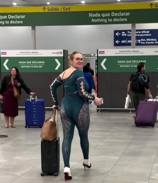 A traveler in a revealing dark green jumpsuit and high heels pulls a suitcase through an airport terminal.