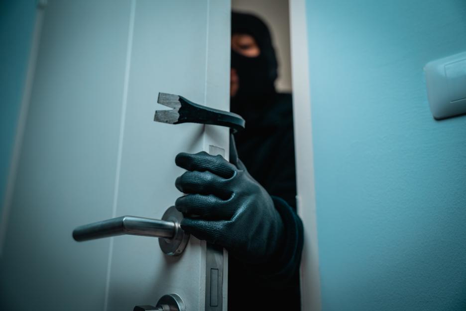 A burglar in a black balaclava, jacket, and gloves uses a crowbar to pry open a white door.