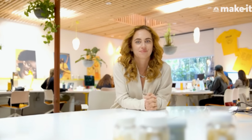 A woman with long curly hair in a light blazer sits at a white table in an office, looking at the camera.