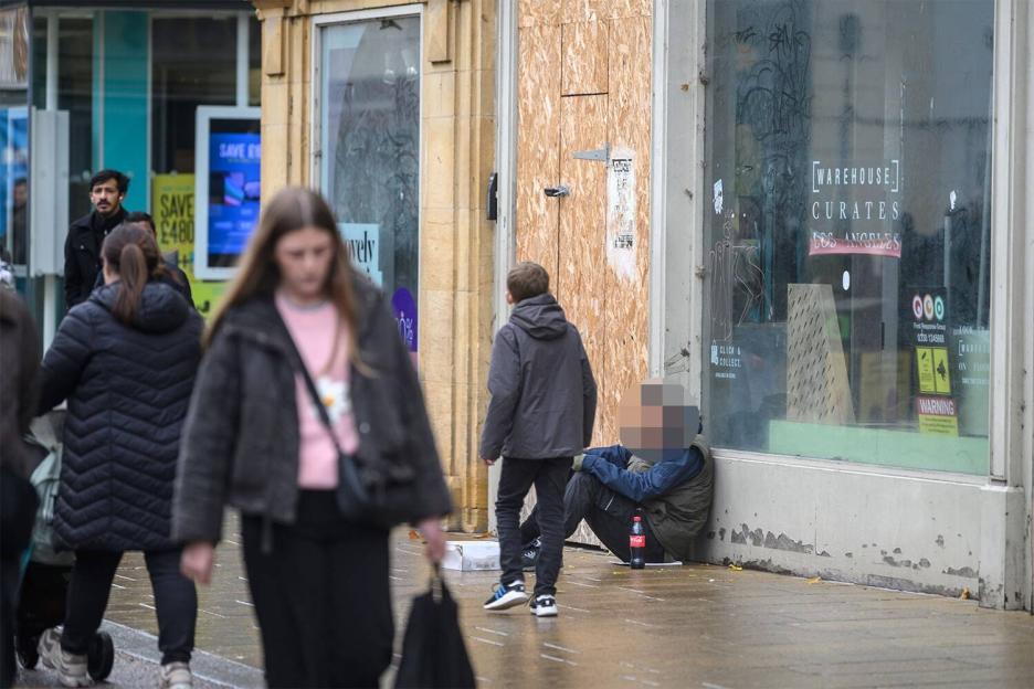 A person sits on the ground next to a boarded-up shop window while people walk by on a wet sidewalk.