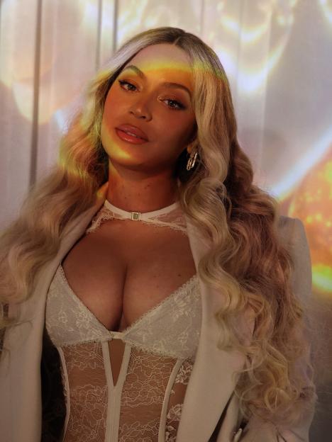 Beyoncé, with blonde wavy hair, wearing a white lace corset and a blazer, poses in front of a warm-toned abstract background.