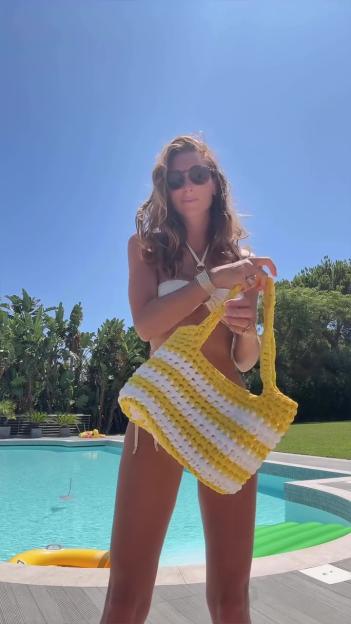 Luisa Zissman displaying her slender frame in a bikini, holding a striped yellow and white crocheted bag by a pool.