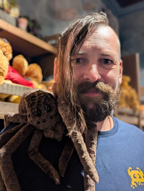 A man with a beard and long hair wearing a blue t-shirt with a plush spider on his shoulder.