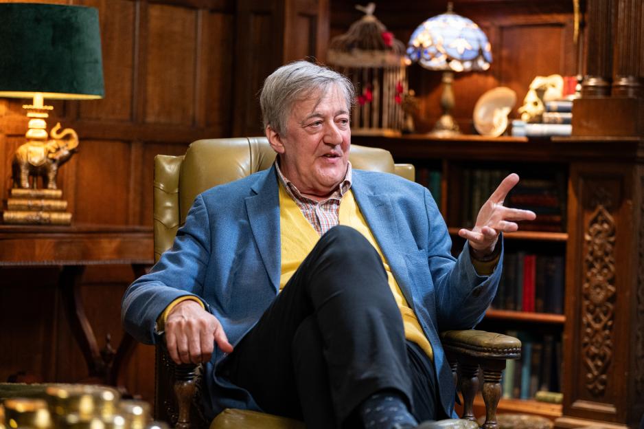 Stephen Fry sitting in a leather chair, wearing a blue jacket and yellow sweater, speaking with an open gesture.
