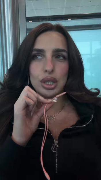 Toni Laites wearing a black top with an open zipper, holding a pink string, and looking away from the camera.