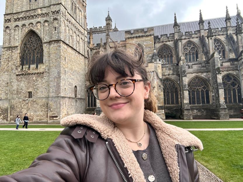 A young woman in a leather and shearling jacket smiling in front of Exeter Cathedral.