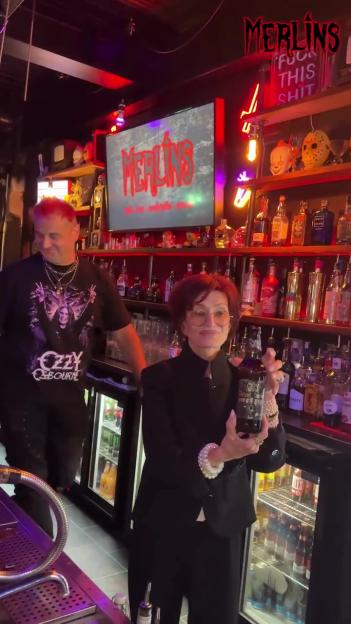 Sharon Osbourne holding a bottle of beer behind a bar, next to a man wearing an Ozzy Osbourne shirt.