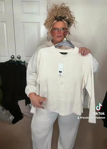 A plus-sized woman in glasses holding up a cream-colored top, wearing cream-colored pants.