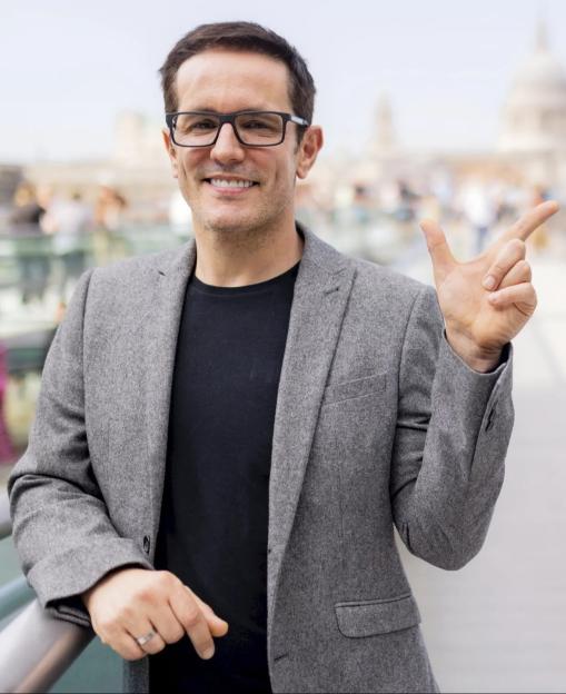 Alastair Wallace, a leadership consultant, smiles at the camera while gesturing with his right hand.