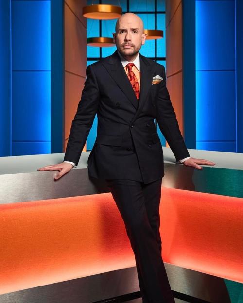 Tom Allen, bald, wearing a dark suit with an orange tie and pocket square, leaning on a counter with an orange glowing panel.