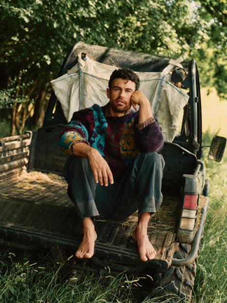 Theo James sitting barefoot in the back of a utility vehicle.