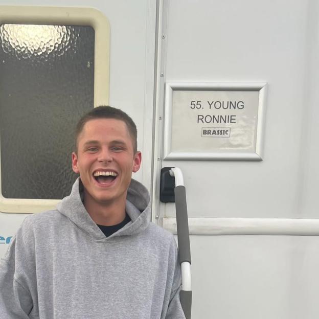 Billy Winter smiling widely in front of a trailer door with a sign that reads "55. YOUNG RONNIE BRASSIC".
