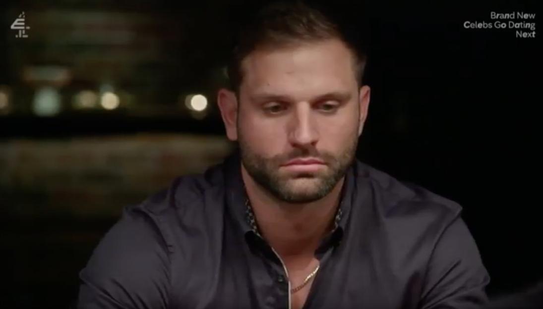 A man with dark hair and a beard, wearing a dark button-down shirt and gold chain, looks down with a somber expression.