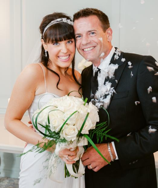 A newlywed couple smiling, the bride holding a white rose bouquet, with confetti falling around them.