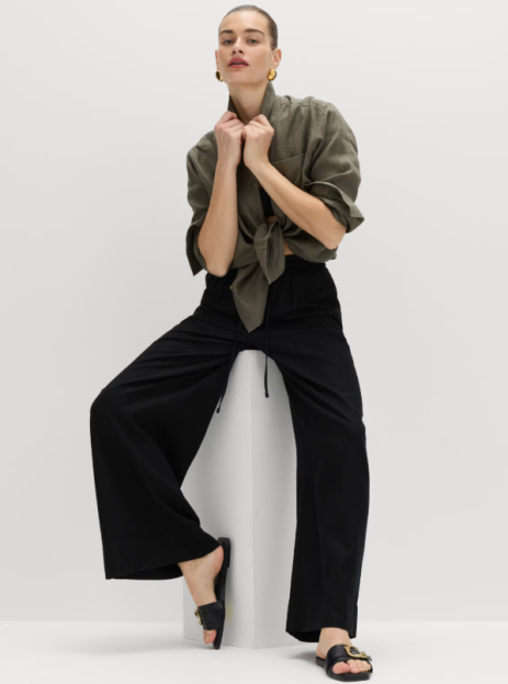 A woman models black linen trousers with a tied olive shirt and black sandals.