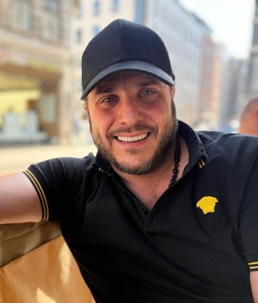 A man wearing a black cap and black polo shirt with a golden Versace logo on it, smiles at the camera.
