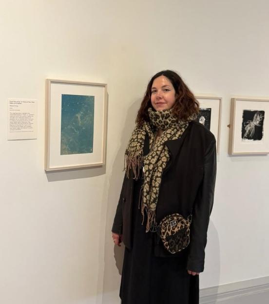 A woman with dark, curly hair wearing a leopard print scarf and black outfit poses in front of framed artwork in an art gallery.