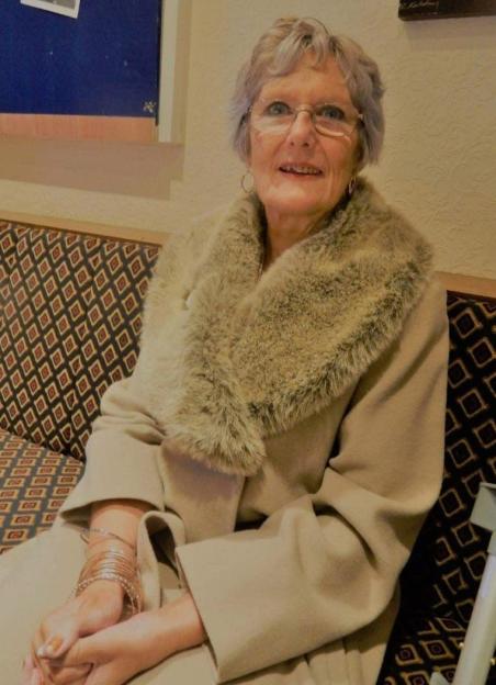 A smiling older woman with grey hair and glasses, wearing a light brown coat with a fur collar, sitting on a patterned couch with her hands folded.
