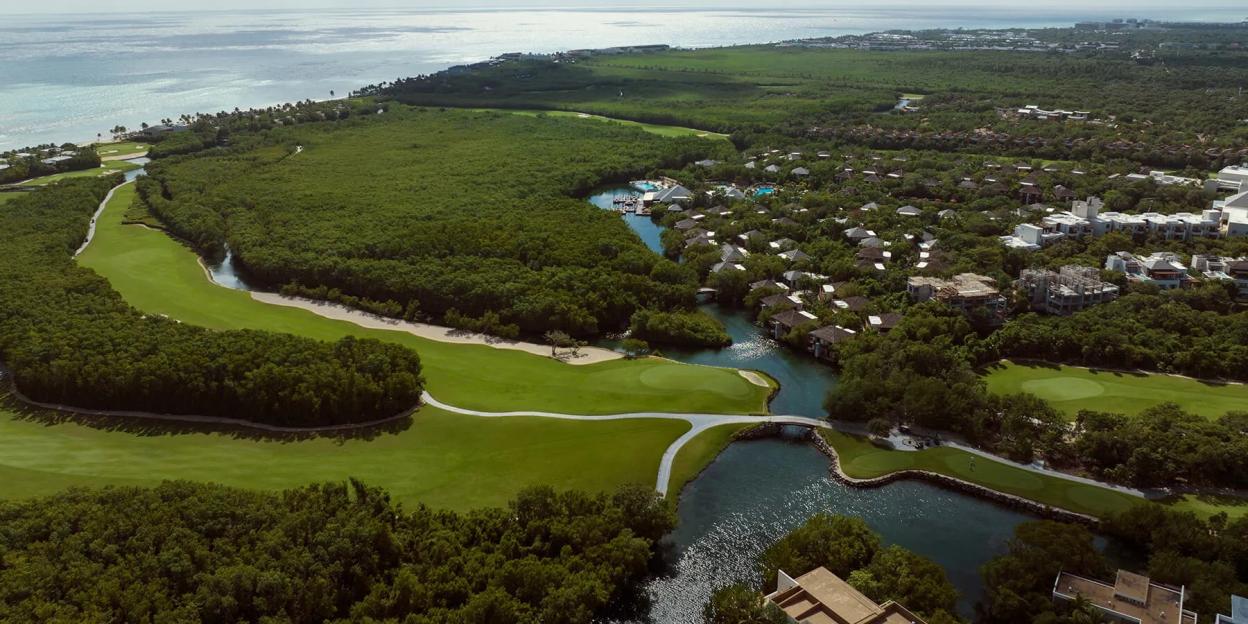 Aerial view of the Fairmount Mayakoba, an award-winning luxury hotel in Riviera Maya, showing a golf course, lush green forest, buildings, and the ocean in the background.