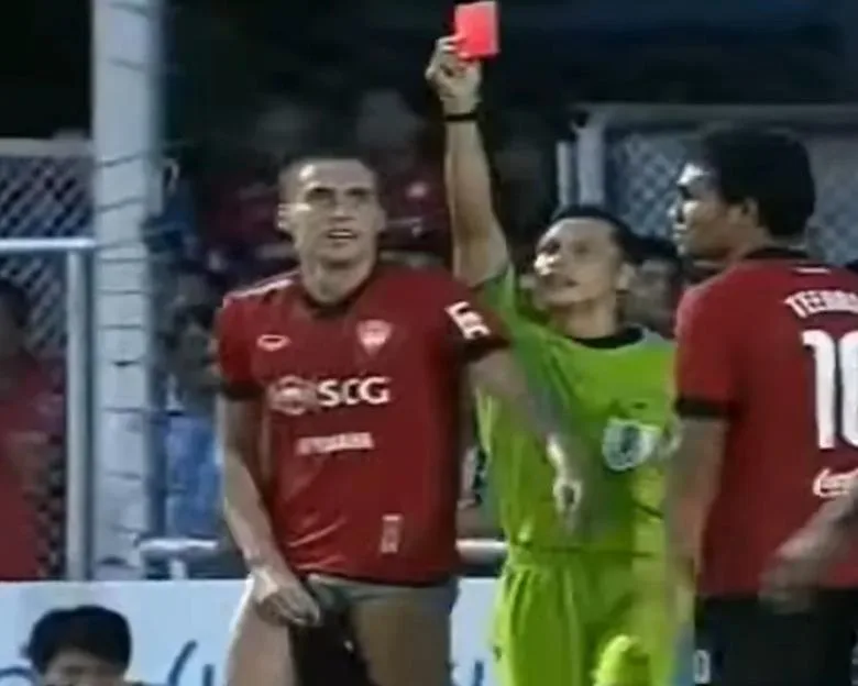 A referee holds up a red card to a football player in a red jersey.
