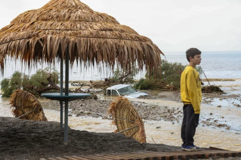 A boy in a yellow sweatshirt stands by a damaged beach bar amid flood debris in Crete.