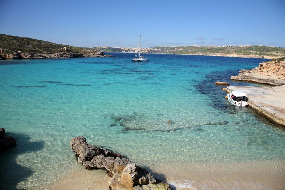 The Blue Lagoon in Gozo, Malta, with clear turquoise water, a yacht, and a small boat docked at a pier.