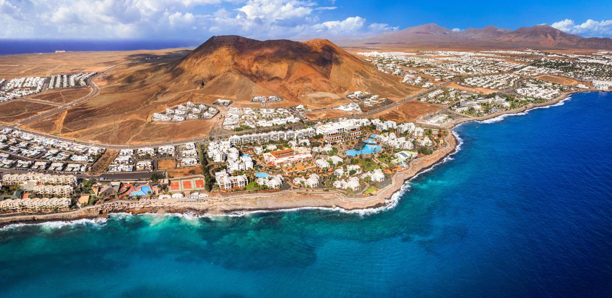 Aerial panoramic view of Playa Blanca resort in Lanzarote, Canary Islands, with a red volcano, ocean, and white buildings.
