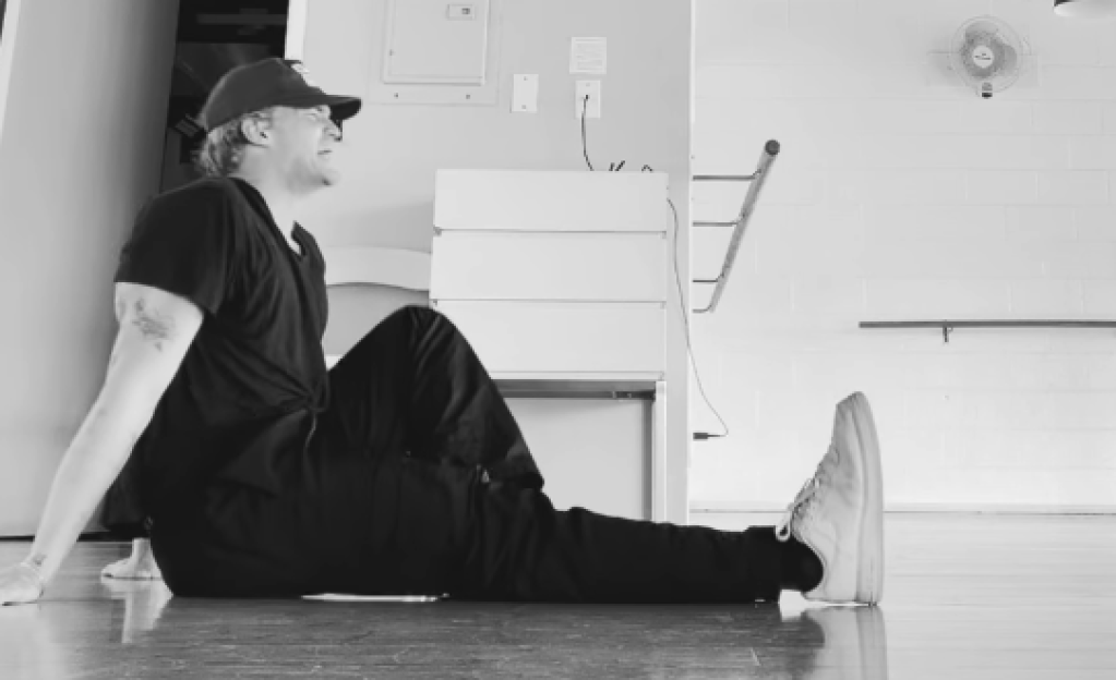 A man in a black shirt and cap sitting on the floor with one leg extended, looking upwards.