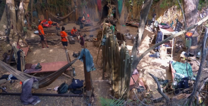 A camp in a jungle with several people relaxing in hammocks and sitting around a small fire.