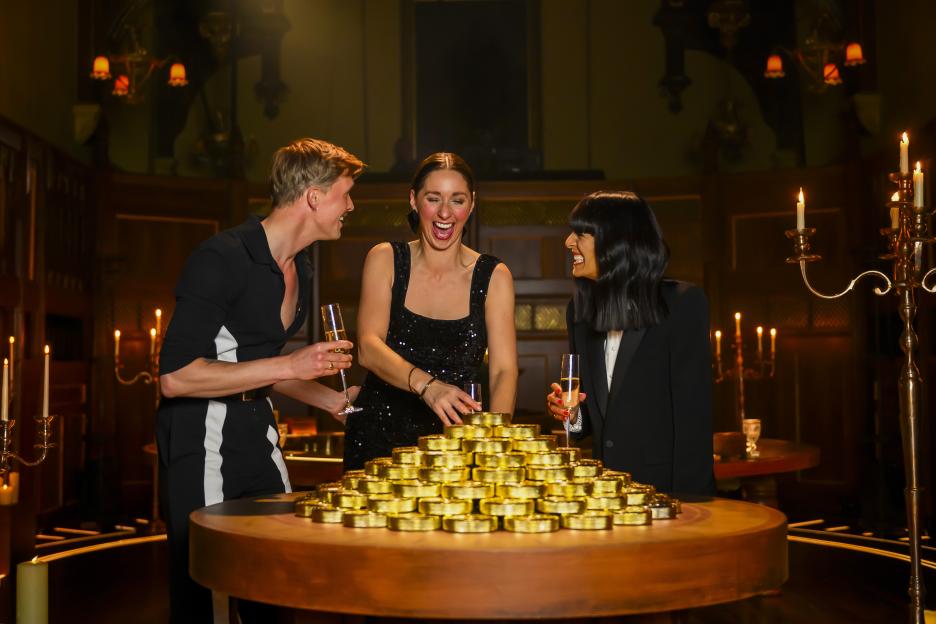 Claudia Winkleman with Stephen and Rachel, winners of season 4 of The Traitors, smiling around a pyramid of golden disks.