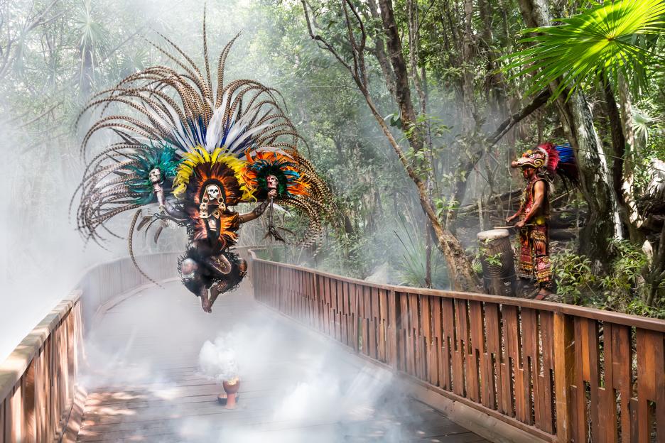 A person in an elaborate feathered costume jumps over a smoking censer on a boardwalk, while another person beats a drum in the jungle.