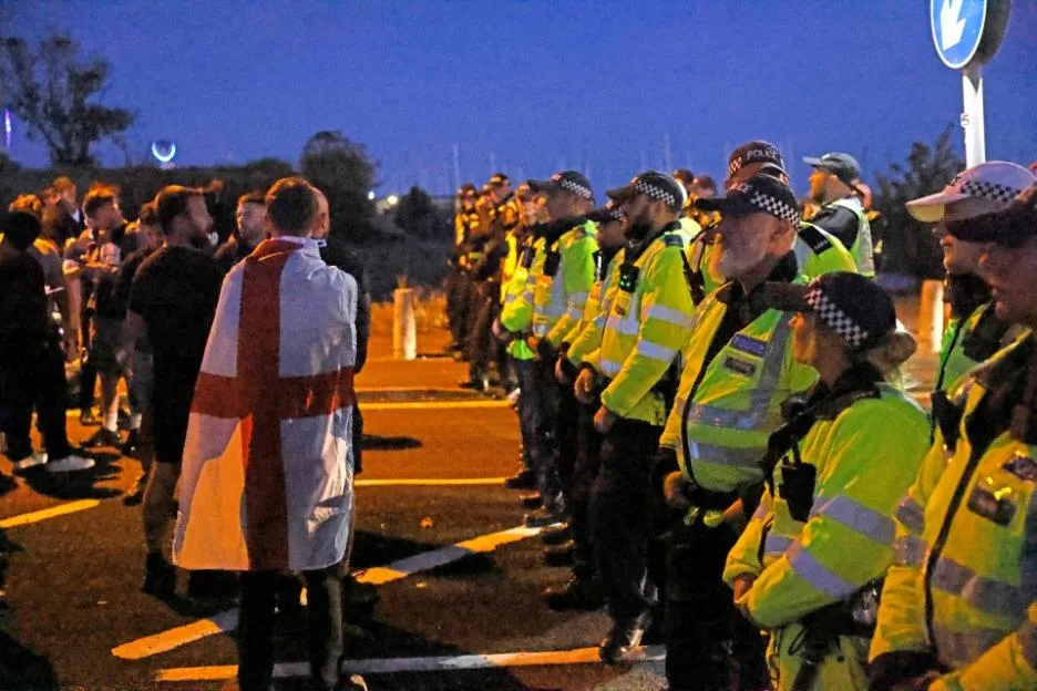 Police line the street as demonstrators hold a protest in Gosport, with a Union flag visible in the foreground.