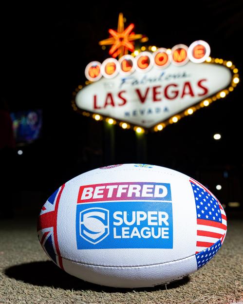 A rugby ball with Betfred Super League logo and flags of the UK and USA on a field, with the Welcome to Las Vegas sign in the background.