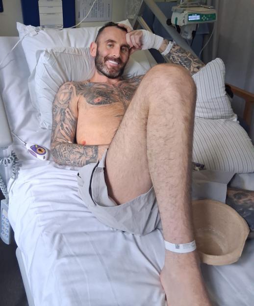 James Bainbridge, diagnosed with stage 4 cancer, lies in a hospital bed with a white wristband on his right ankle, an IV drip visible behind him.