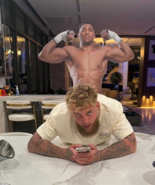 Jake Paul lying on a table in front of Anthony Joshua, who is flexing.