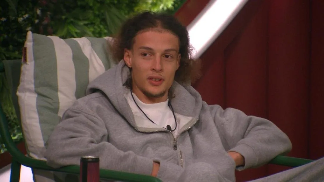 A young man with curly dark hair wearing a gray zip-up hoodie is seated in a striped chair, looking off to the side.