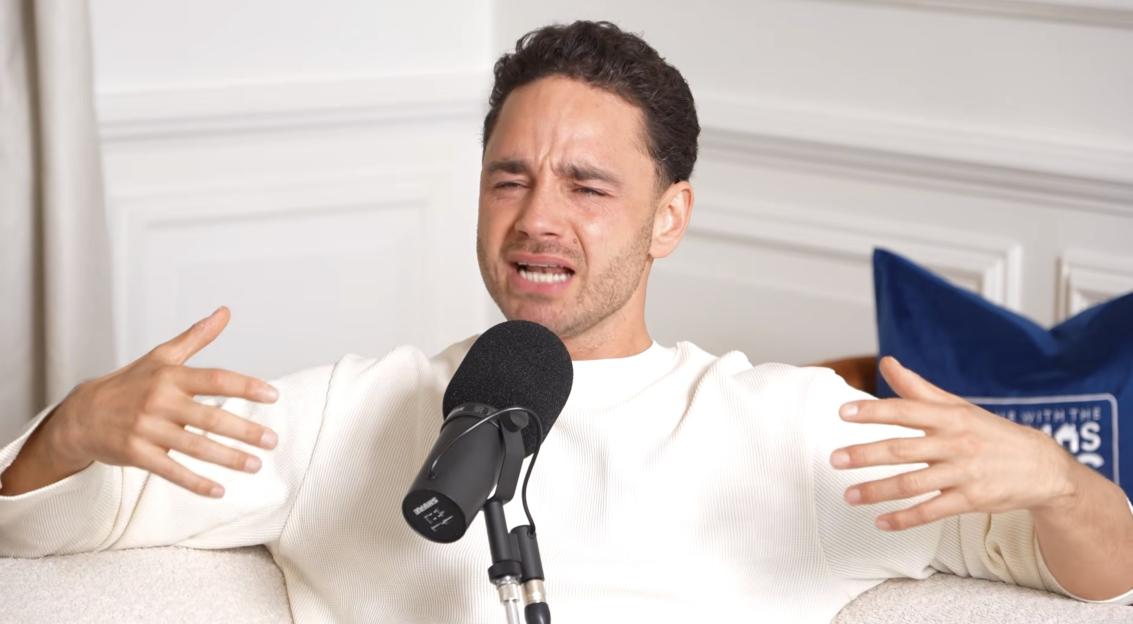 Adam Thomas sitting on a couch and speaking into a microphone with his hands gesturing.