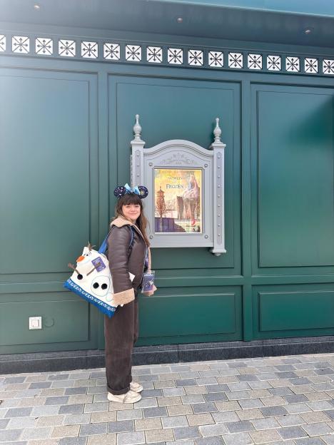 A young woman wearing Mickey Mouse ears and an Olaf backpack standing in front of a "World of Frozen" preview poster.