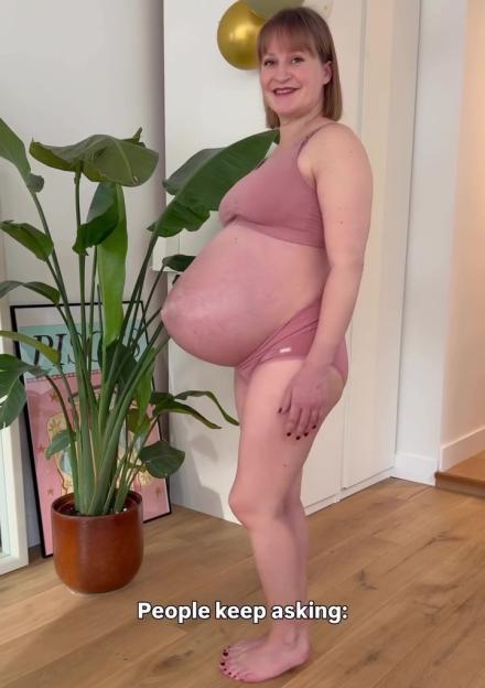 A pregnant woman expecting triplets displays her large baby bump while wearing a pink bra and underwear.