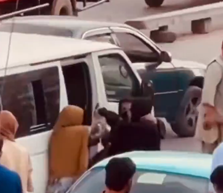 A woman in a brown hijab is being forcefully pulled from a white van by a person in dark clothing, while other people and vehicles are visible in the background.
