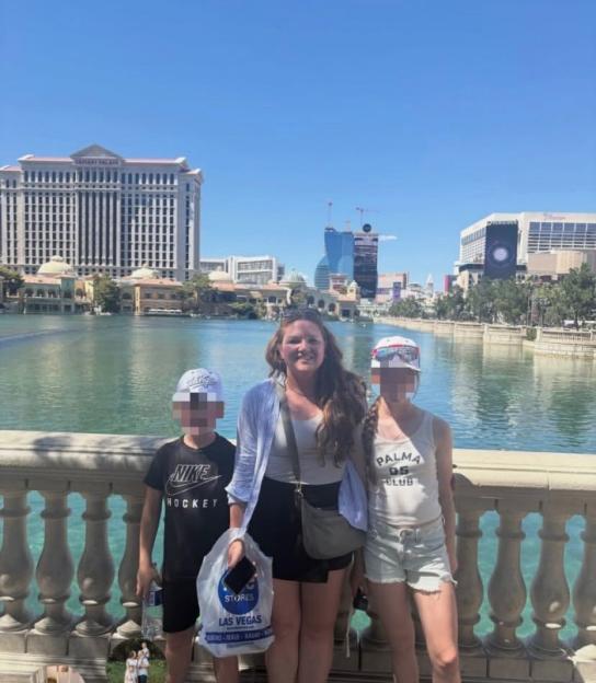 A woman and two children with their faces blurred, standing in front of a body of water with buildings in the background.