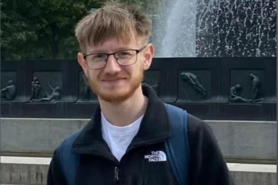 A young man with glasses and light brown hair and beard smiles in front of a fountain.