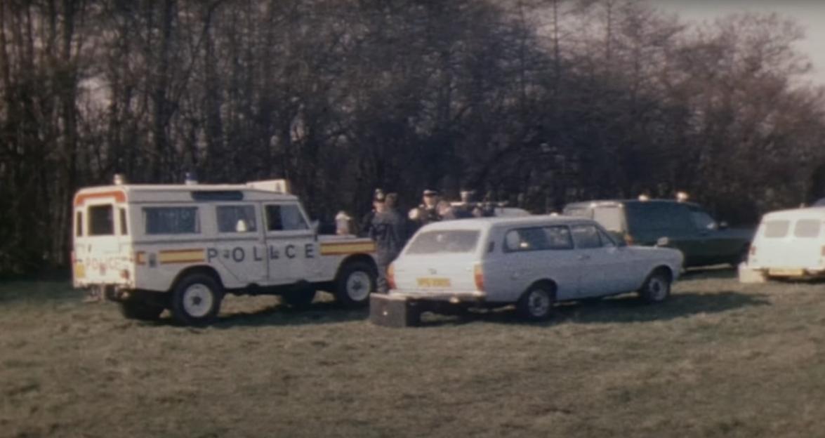 Police vehicles and officers in a field, with trees in the background.