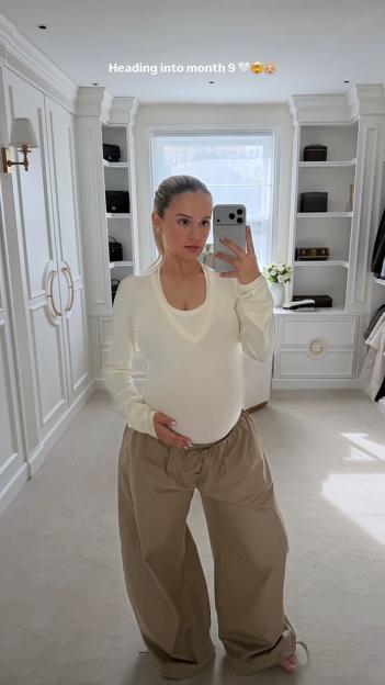 Pregnant woman in an off-white long-sleeved shirt and loose khaki pants taking a mirror selfie.