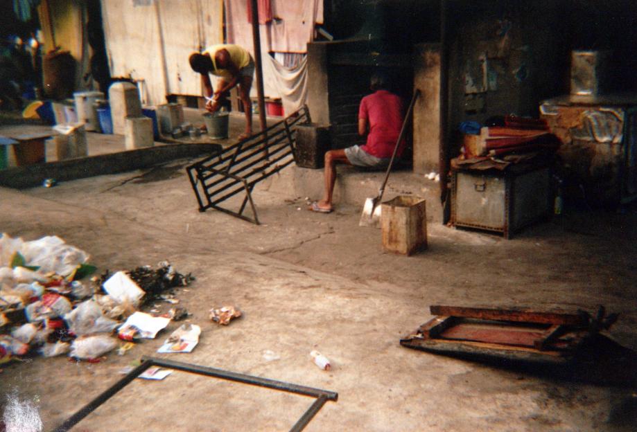 A man in a yellow tank top washes items in a bucket while another man in a red shirt and shorts sits with a shovel.