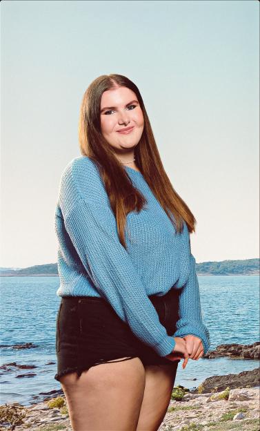 A smiling young woman in a blue sweater and black shorts poses in front of a body of water.