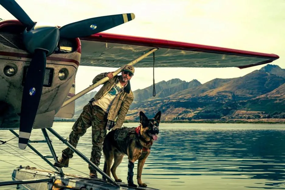 Brad Pitt stands next to a combat dog on the float of a seaplane parked by a lake, with mountains in the background.