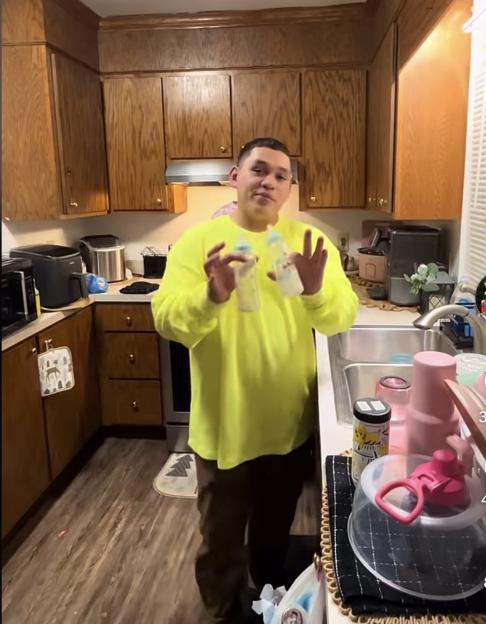 A man in a neon yellow shirt stands in a kitchen, holding two baby bottles.