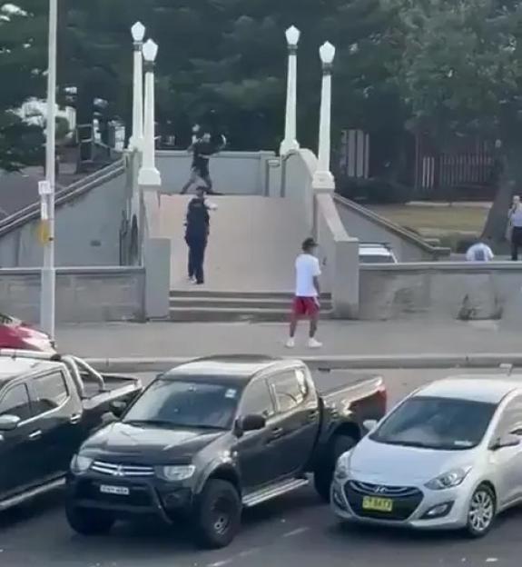A man appearing to be shot at and assaulted by another civilian on a bridge, then cuffed by police, with cars parked in the foreground.