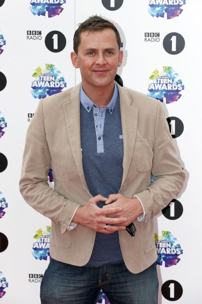 Radio 1 DJ Scott Mills on a red carpet with "BBC Radio 1 Teen Awards" branding in the background.